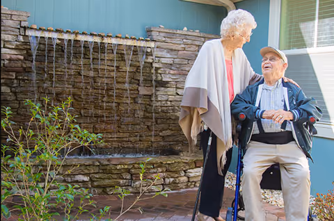 An elderly woman standing and smiling at an elderly man seated in a wheelchair outside near a stone water fountain wall with water flowing from multiple spouts. There are plants and a building with windows in the background.