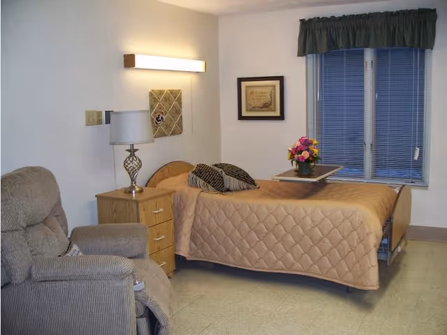 A tidy senior bedroom with a single bed covered by a beige quilt, bedside table with lamp, recliner chair, window with blinds, and flowers on an overbed table.