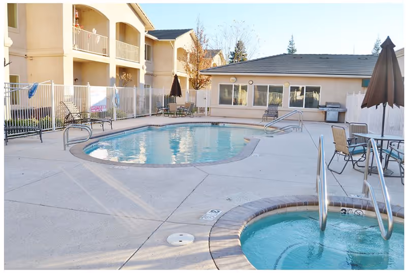 Outdoor swimming pool and hot tub area surrounded by patio furniture and umbrellas, with a beige building and fenced area in the background under clear daylight.