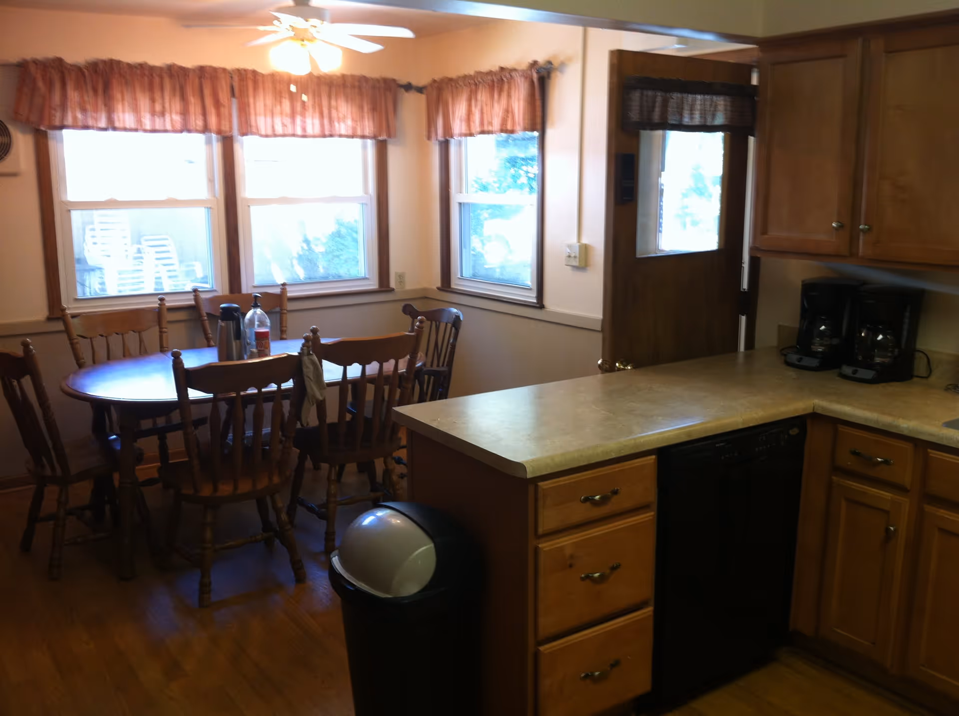 Sunlit kitchen and dining area with wooden cabinets, a countertop with coffee makers, and a round dining table with chairs.
