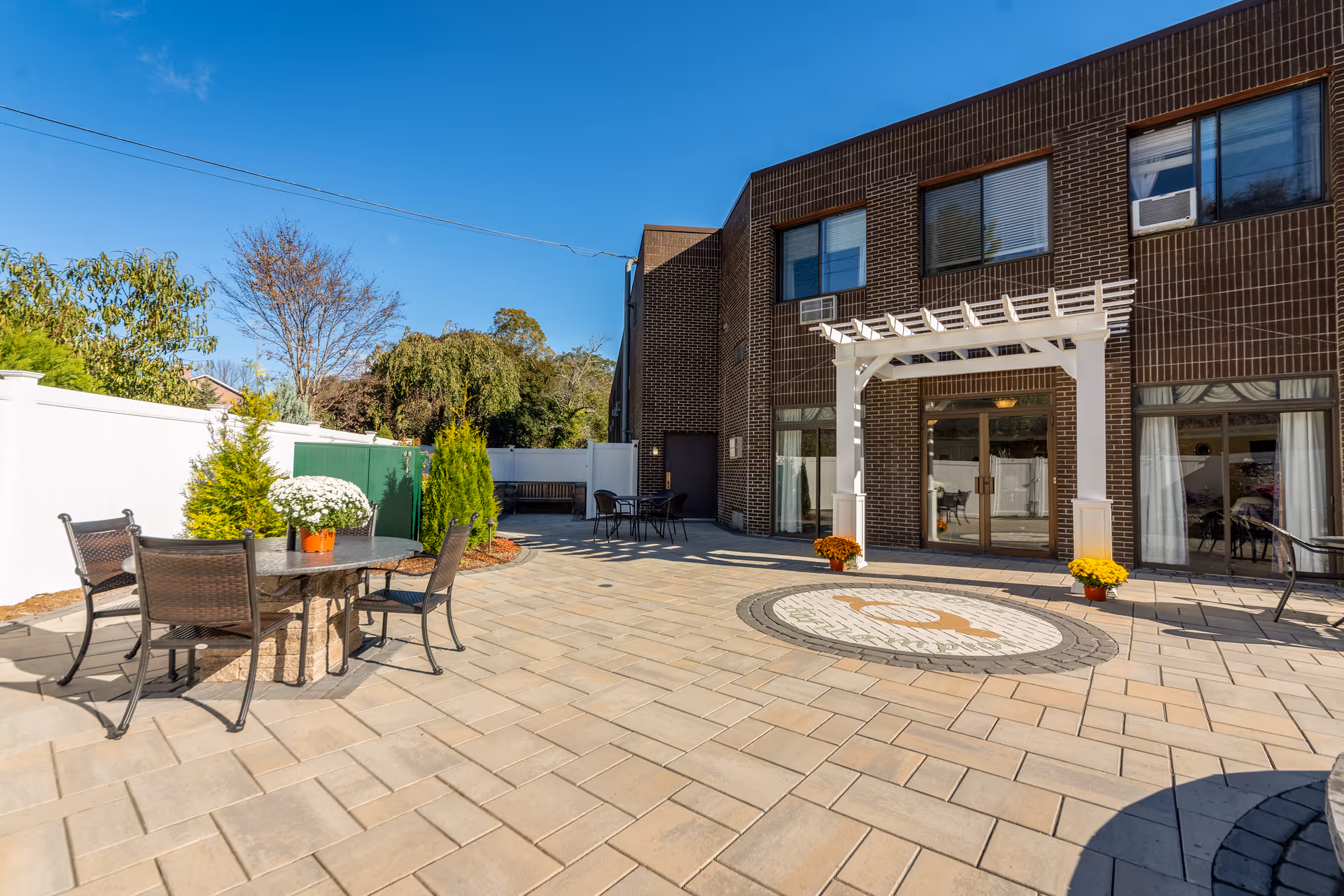 Outdoor patio area at CareOne at Northampton with a paved stone floor, a round table with four chairs, potted plants, and a white pergola attached to a brick building under a clear blue sky.