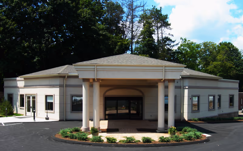 Front entrance of a single-story care facility with a covered porte-cochere supported by columns and a circular driveway.
