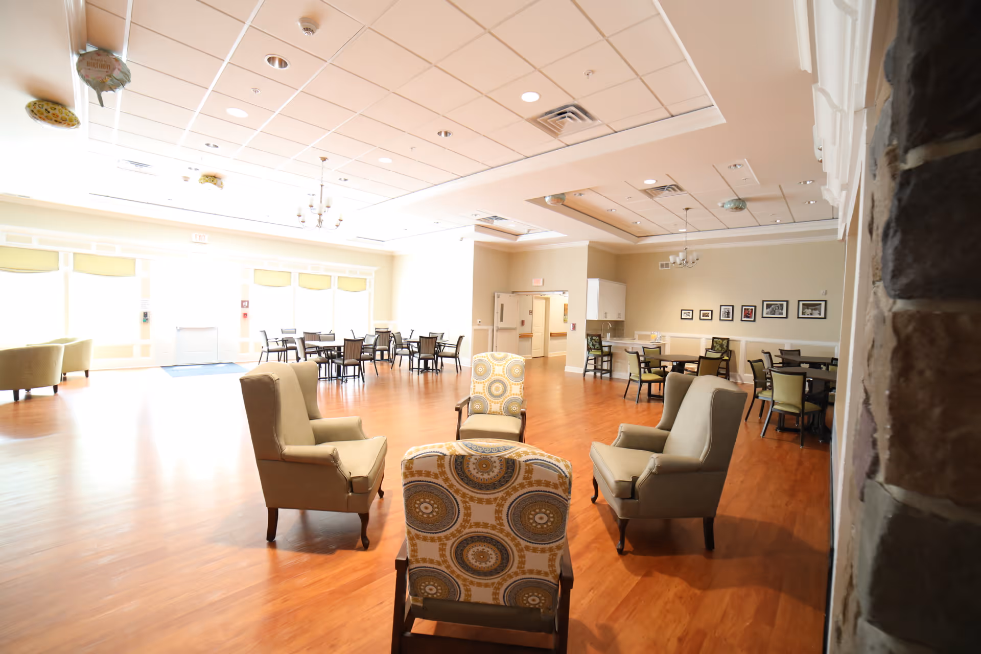 A spacious, well-lit common area with wooden flooring featuring several armchairs arranged in a circle in the foreground and multiple dining tables with chairs in the background. The room has a high ceiling with recessed lighting and chandeliers, and large windows letting in natural light.