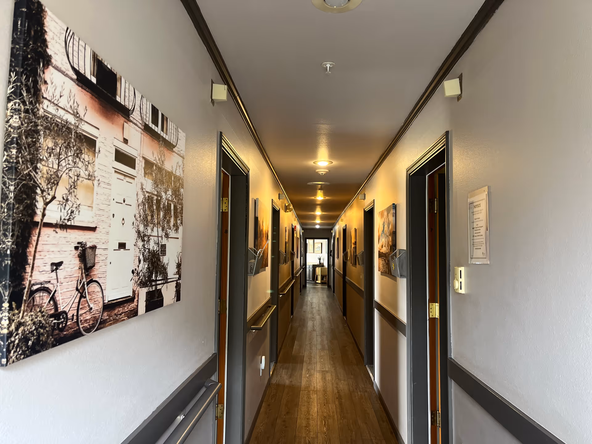 A long, narrow hallway in an assisted living facility with wooden flooring and light gray walls. Several doors line both sides of the hallway, and framed pictures hang on the walls. The hallway is lit by ceiling lights, and there is a window at the far end letting in natural light.