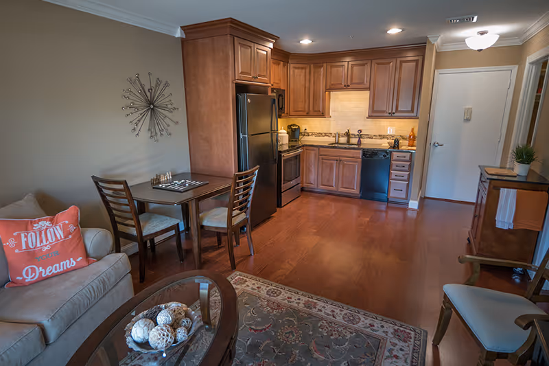 Interior view of a senior living facility unit featuring a small kitchen with wooden cabinets, black appliances including a refrigerator, stove, and dishwasher, a dining table with two chairs, a beige couch with a decorative pillow that says 'Follow your Dreams', a coffee table with decorative items, and a patterned rug on wooden flooring.
