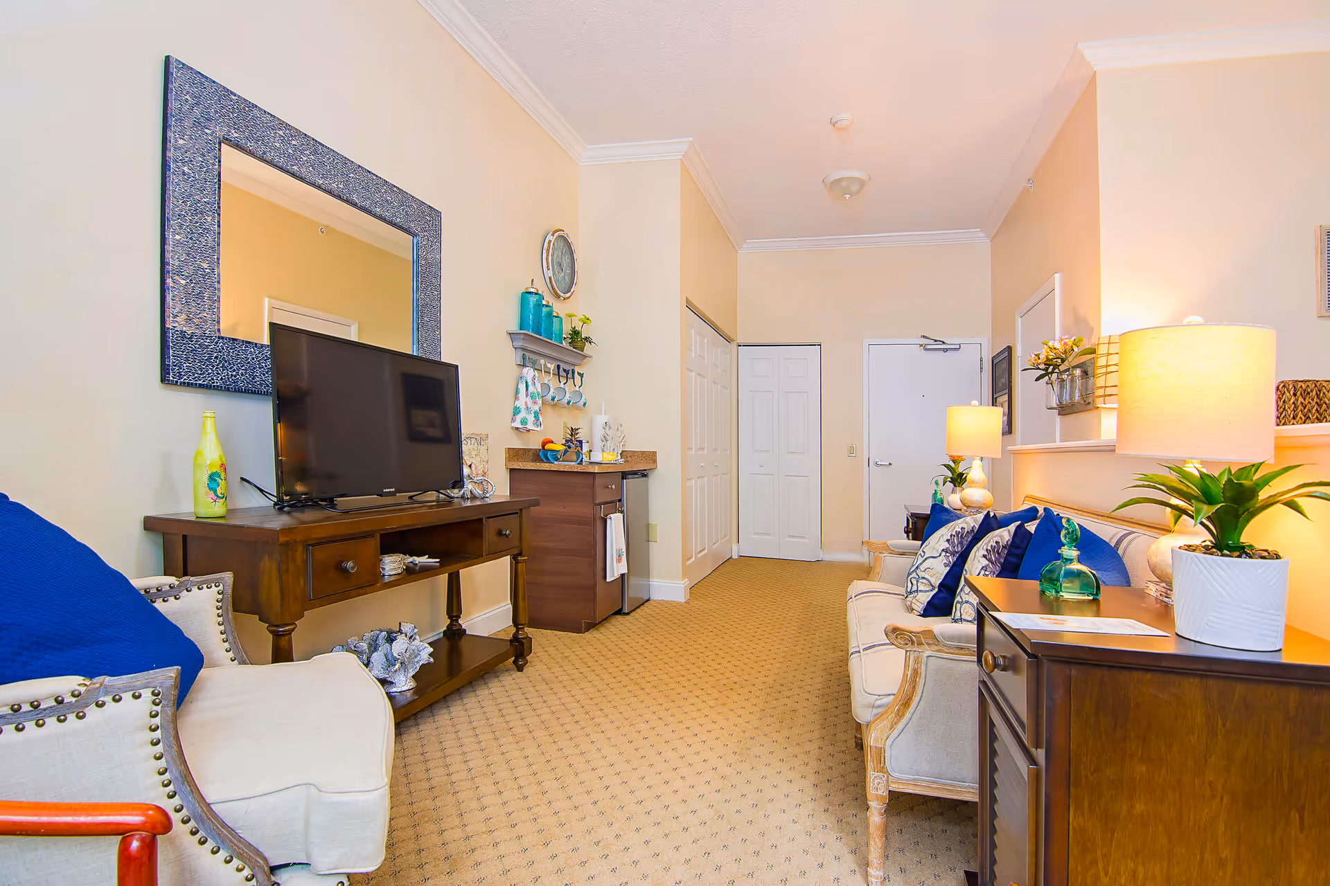 A cozy living room area in a senior living facility featuring a beige armchair with a blue cushion, a wooden TV stand with a flat-screen television, a large decorative mirror on the wall, and a small kitchenette area with shelves holding cups and decorative items. Opposite the TV stand is a beige sofa with blue and patterned cushions, a wooden side table with a lamp and a potted plant, and another lamp on a small table near the entrance door. The room has beige walls and carpeted flooring.