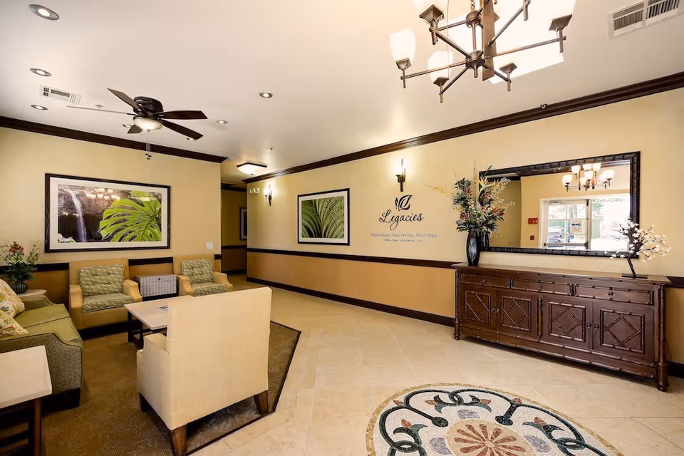 A cozy interior lounge area with beige walls and dark wood trim. The room features a ceiling fan, a chandelier, framed botanical artwork, a large mirror above a wooden sideboard with decorative flowers, and comfortable seating including armchairs and a sofa arranged around a coffee table.