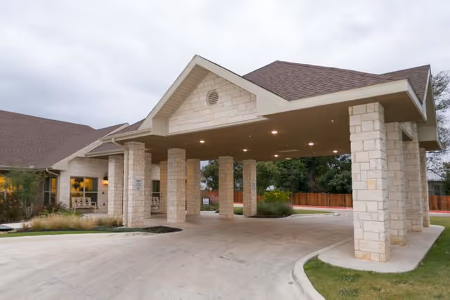 Covered entrance area of a healthcare and rehabilitation facility with stone pillars supporting a peaked roof, surrounded by a driveway and landscaped greenery under an overcast sky.