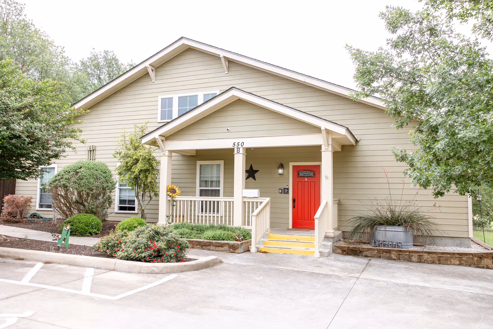 Exterior view of a single-story building with beige siding and a red front door under a small covered porch. The building is surrounded by landscaped bushes, trees, and flowers, with a concrete driveway and parking area in front.