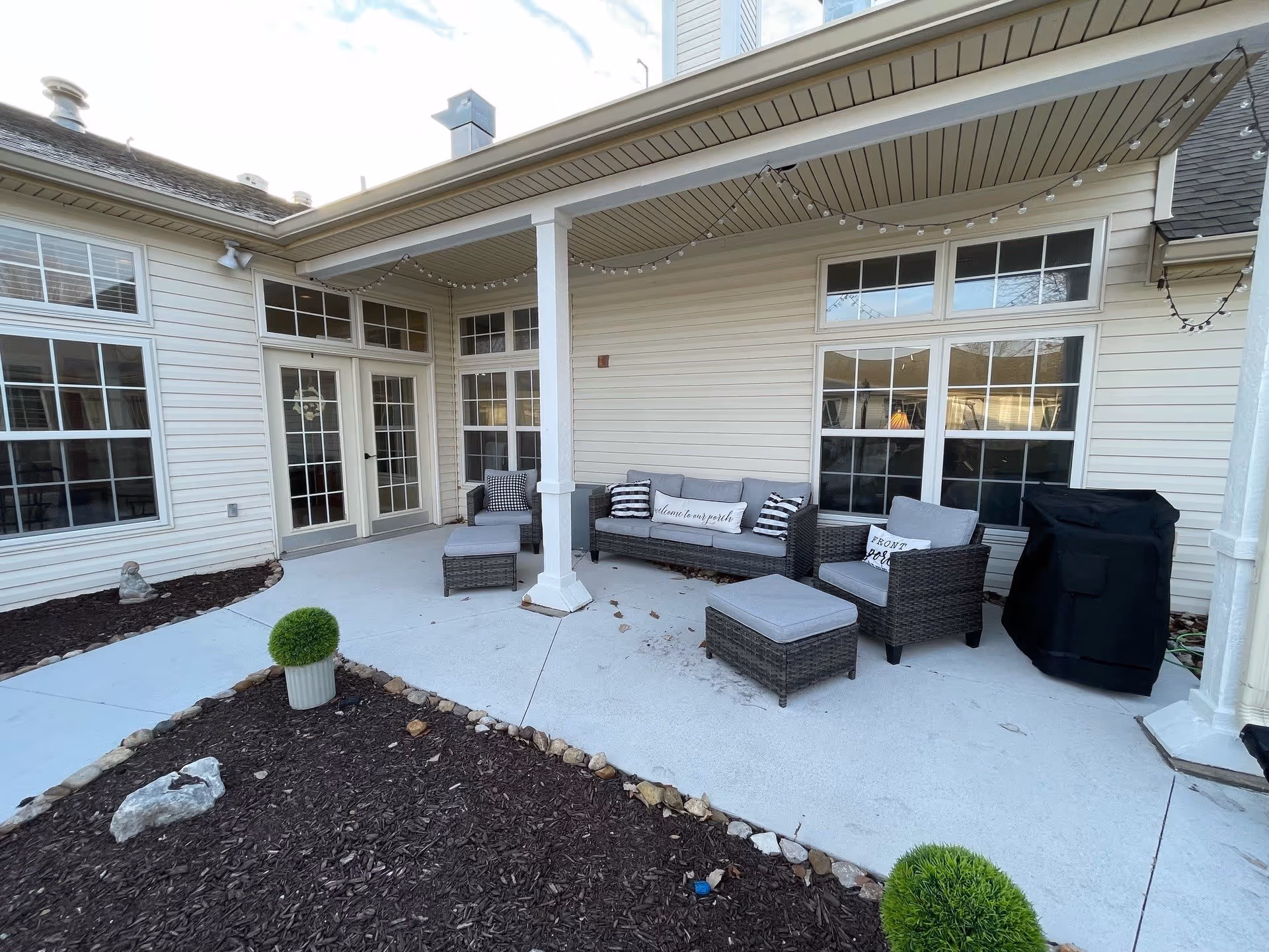 Outdoor covered patio area with cushioned wicker furniture including a sofa, two armchairs, and two ottomans. The patio is adjacent to a building with large windows and glass doors. There are string lights hanging from the ceiling and a covered grill on the right side. Mulched garden beds with small green shrubs border the patio.
