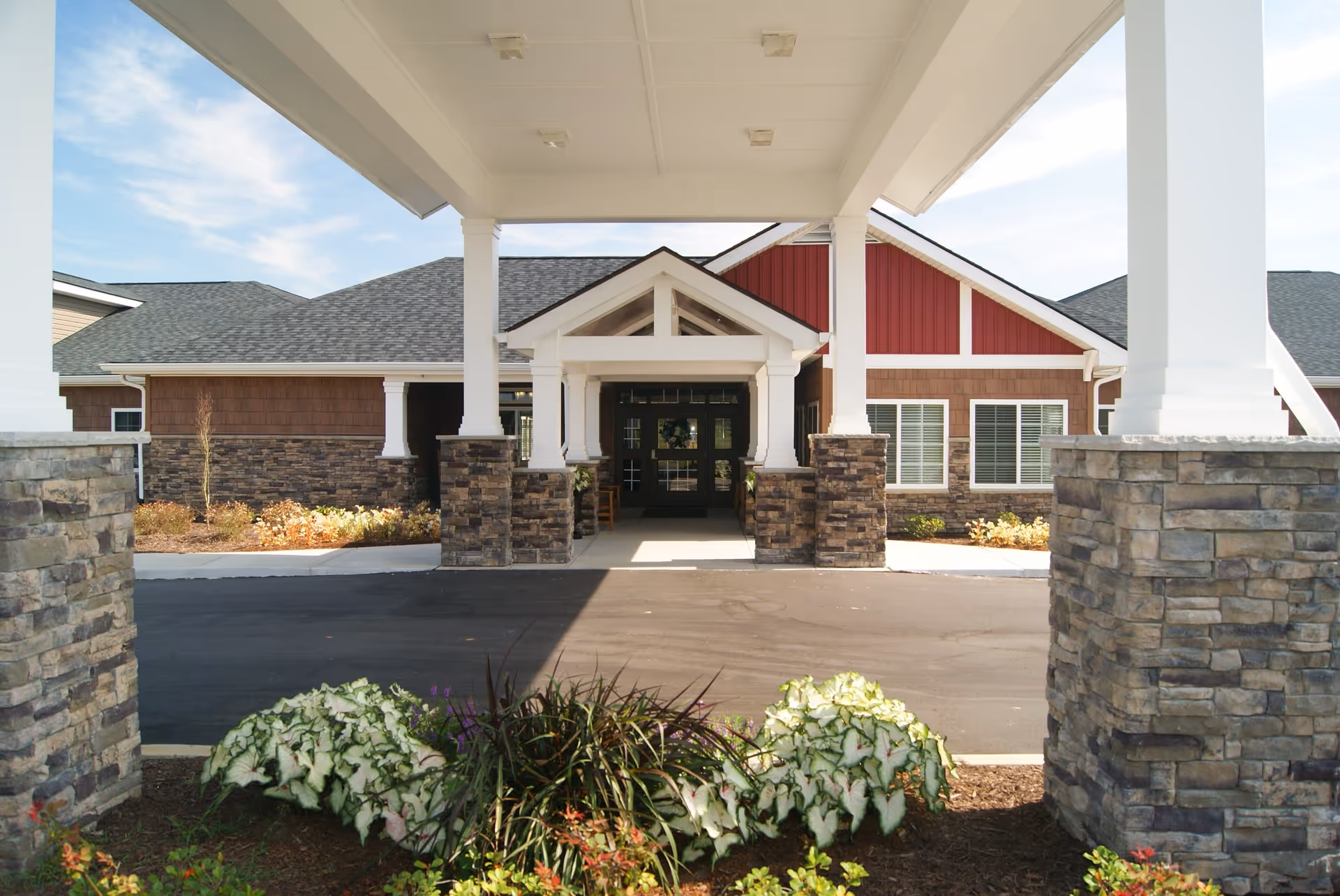 Entrance of a senior living facility with a covered driveway supported by white pillars with stone bases. The building has a combination of red and brown siding with white trim and multiple windows. There are landscaped plants and flowers in front of the entrance.