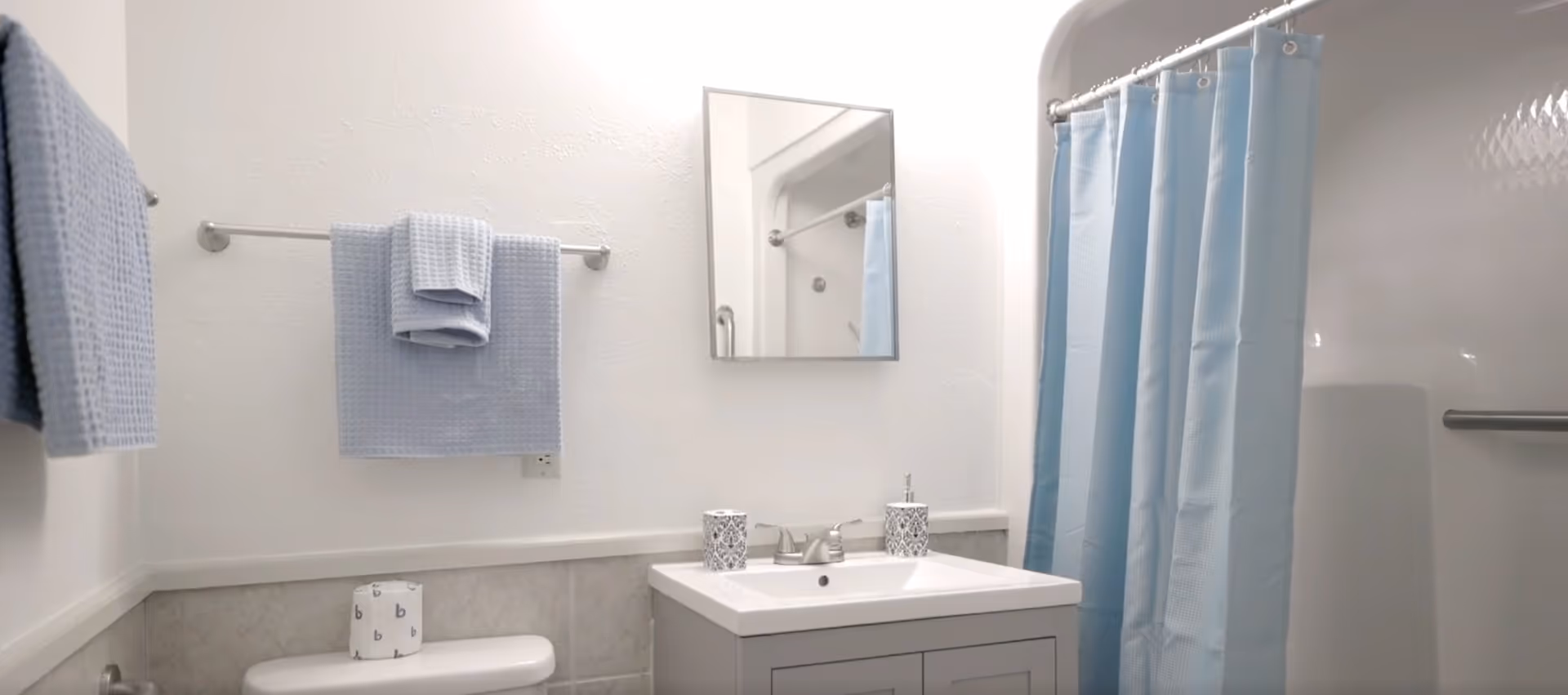 Small bright bathroom featuring a sink and vanity with mirror, toilet, towel rack with blue towels, and a blue shower curtain.