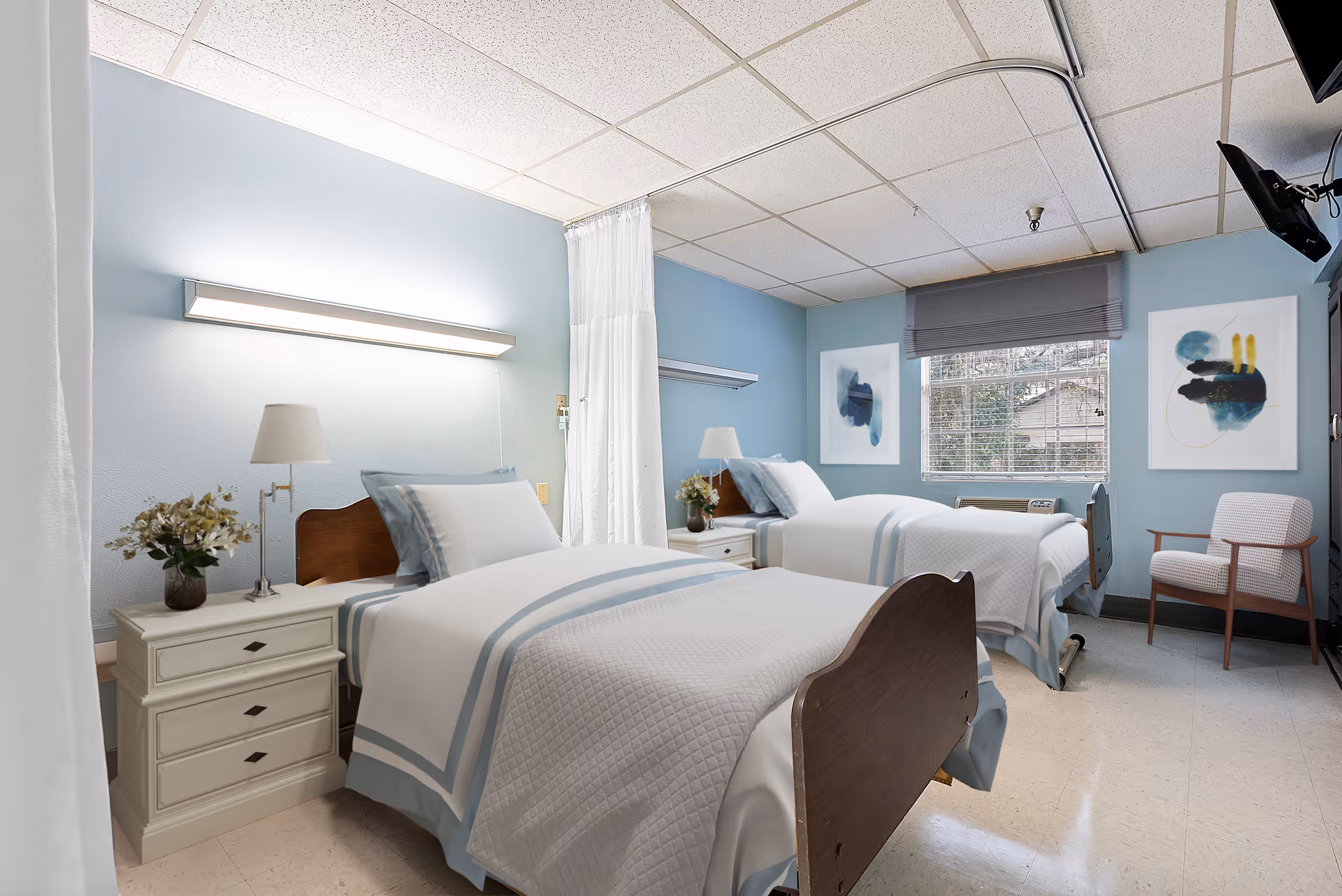 Shared patient bedroom with two single beds, bedside tables and lamps, artwork, a window, and a chair.