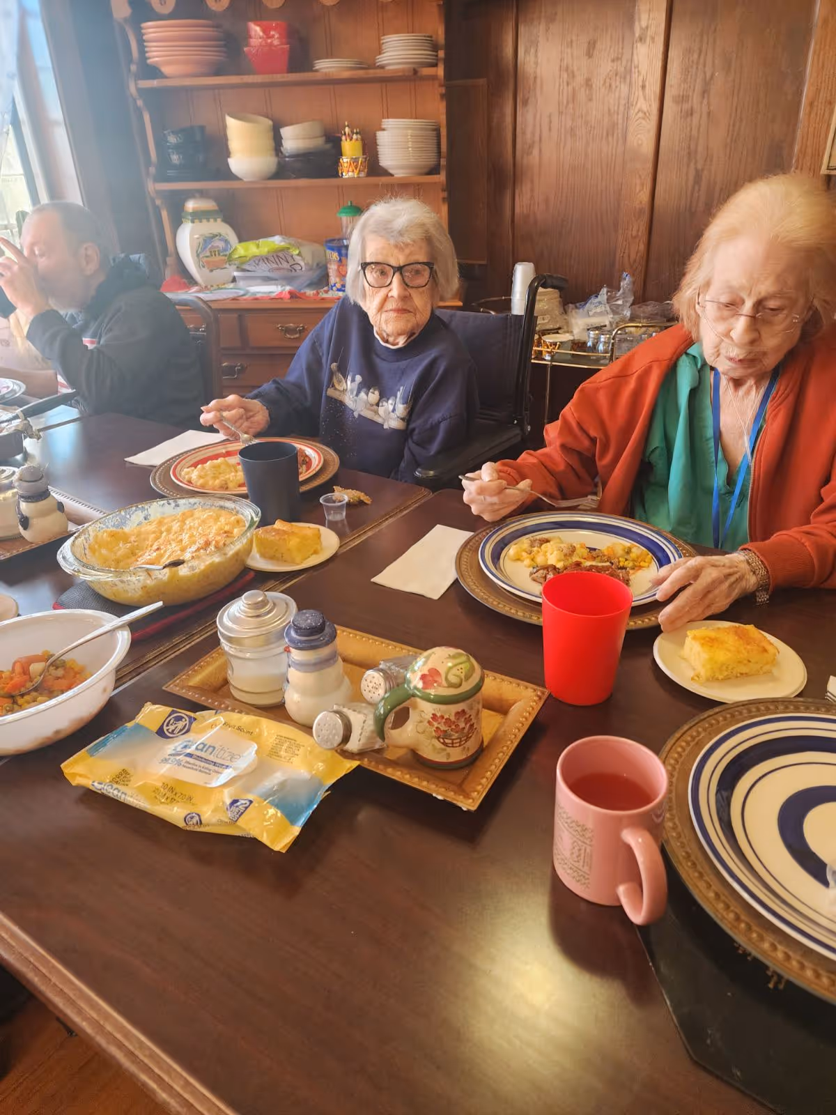 Elderly residents eating a meal at a communal dining table with plates, bowls, cups, and serving dishes in a wood-paneled room.