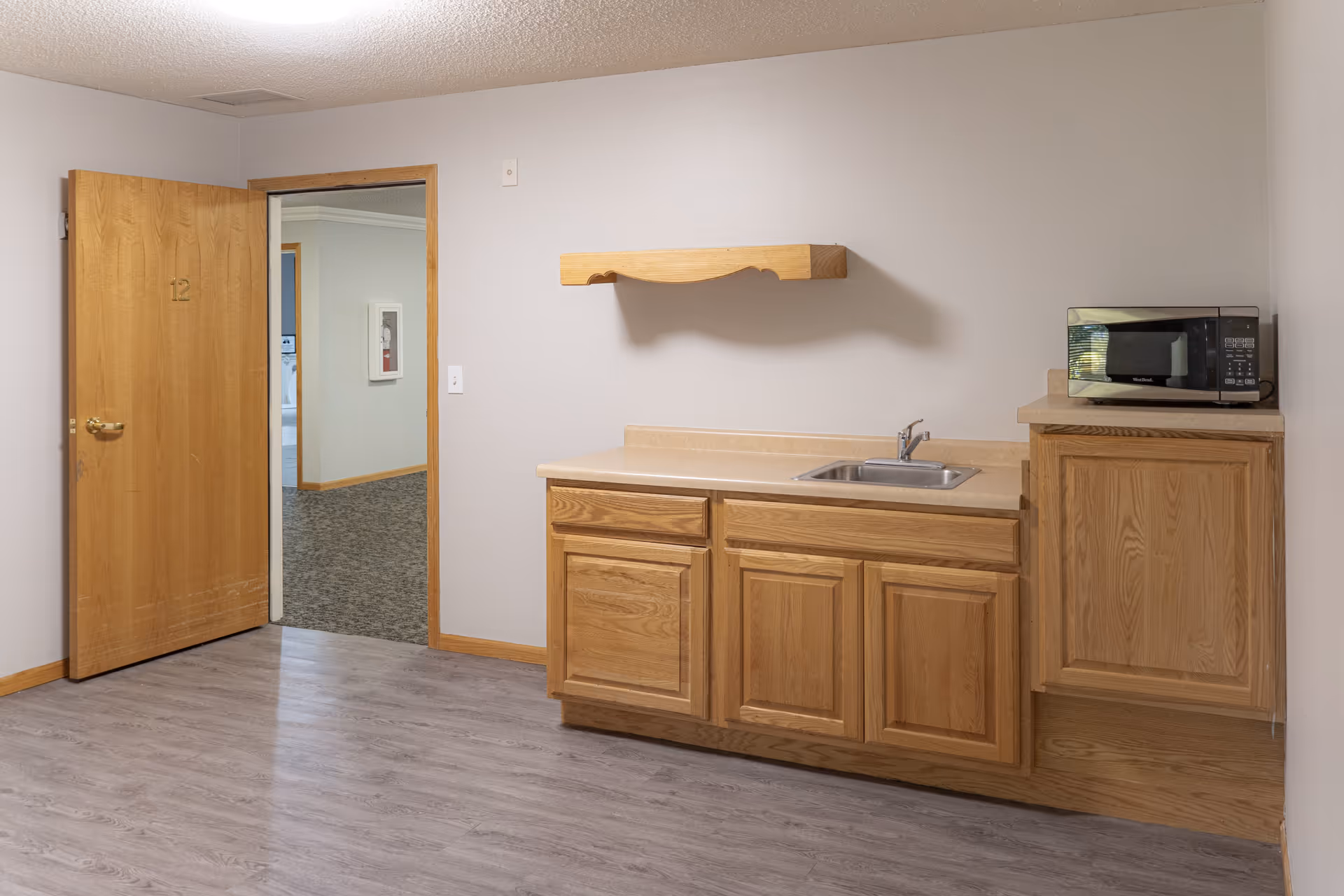 A small kitchenette area with wooden cabinets, a countertop with a sink, and a microwave on the right side. The room has light gray walls and wood-look flooring. An open wooden door with the number 12 leads to a hallway with carpeted flooring and a fire extinguisher cabinet on the wall.