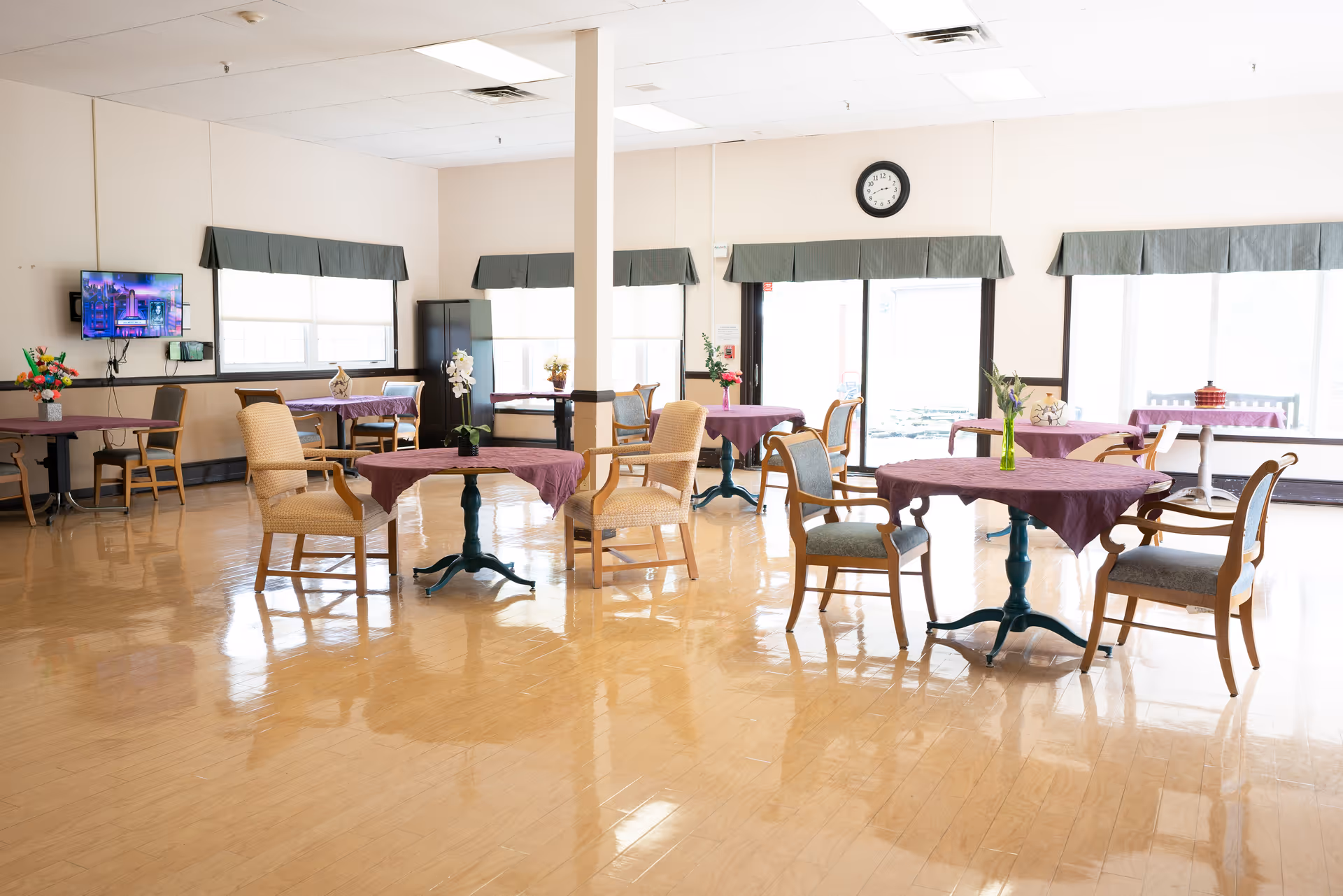 A bright dining room with several round tables covered with purple tablecloths, each surrounded by wooden chairs with cushions. The room has large windows with green valances, a wall-mounted TV, and a shiny wooden floor. There are flower arrangements on the tables and a door leading outside.