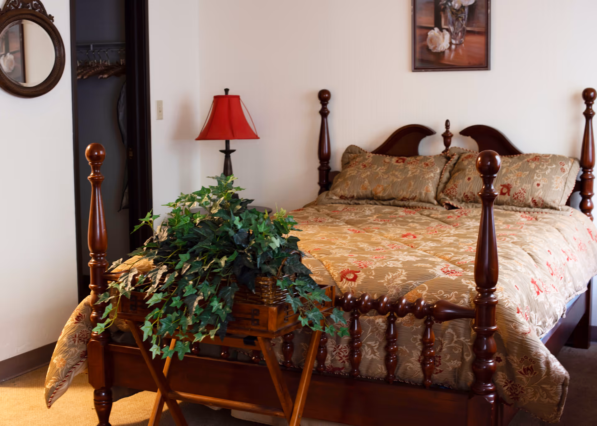 A bedroom with a wooden four-poster bed covered with a floral patterned bedspread and matching pillows. A wooden tray table at the foot of the bed holds a lush green plant. To the left, there is a red lampshade on a black lamp base and an open closet door with hangers inside. A round mirror hangs on the wall near the closet, and a framed floral picture is mounted above the bed.