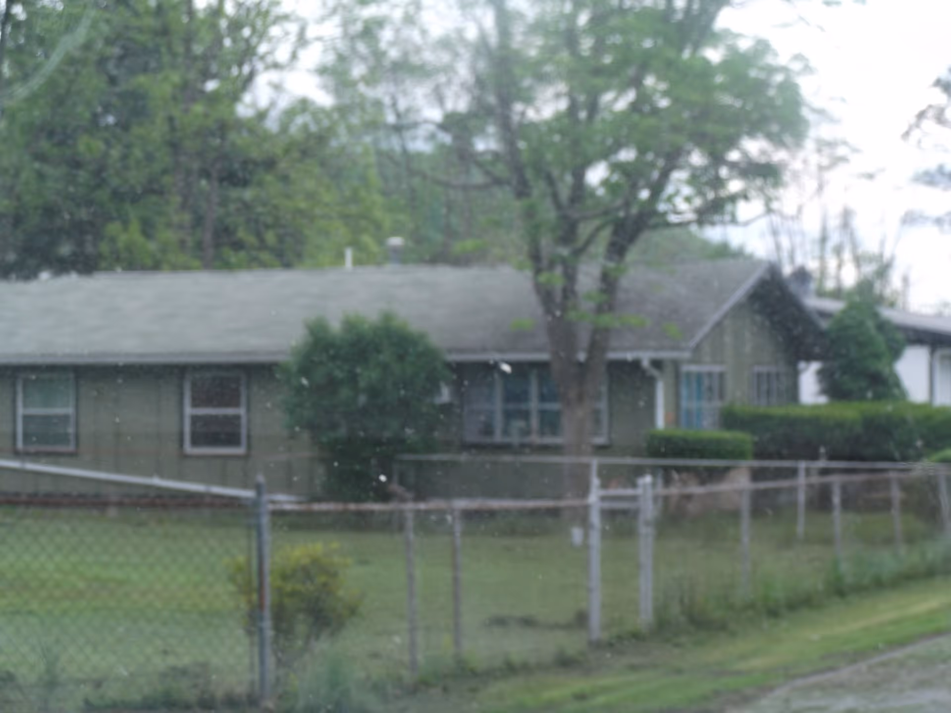 A single-story building with a gray roof and green exterior walls, surrounded by trees and bushes. There is a chain-link fence in the foreground and a grassy area between the fence and the building.