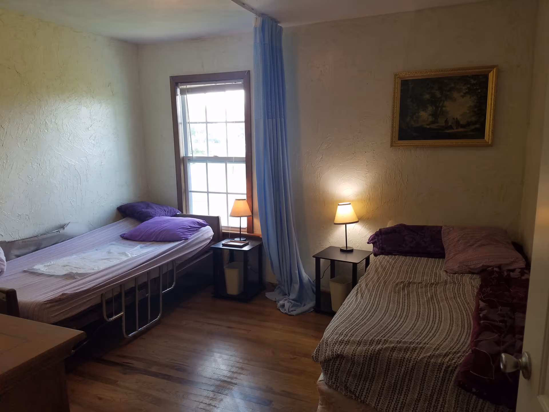 A small bedroom with two single beds placed against opposite walls. Each bed has pillows and blankets, with a small nightstand and a lamp beside each bed. There is a window with a blue curtain in the middle of the room, and a framed painting hangs on the wall above one of the beds. The floor is wooden, and the walls have a textured finish.