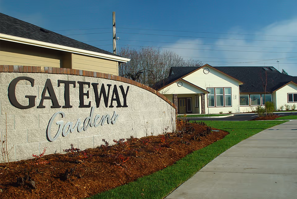 Stone entrance sign reading 'GATEWAY Gardens' with landscaped beds and a single-story building in the background.