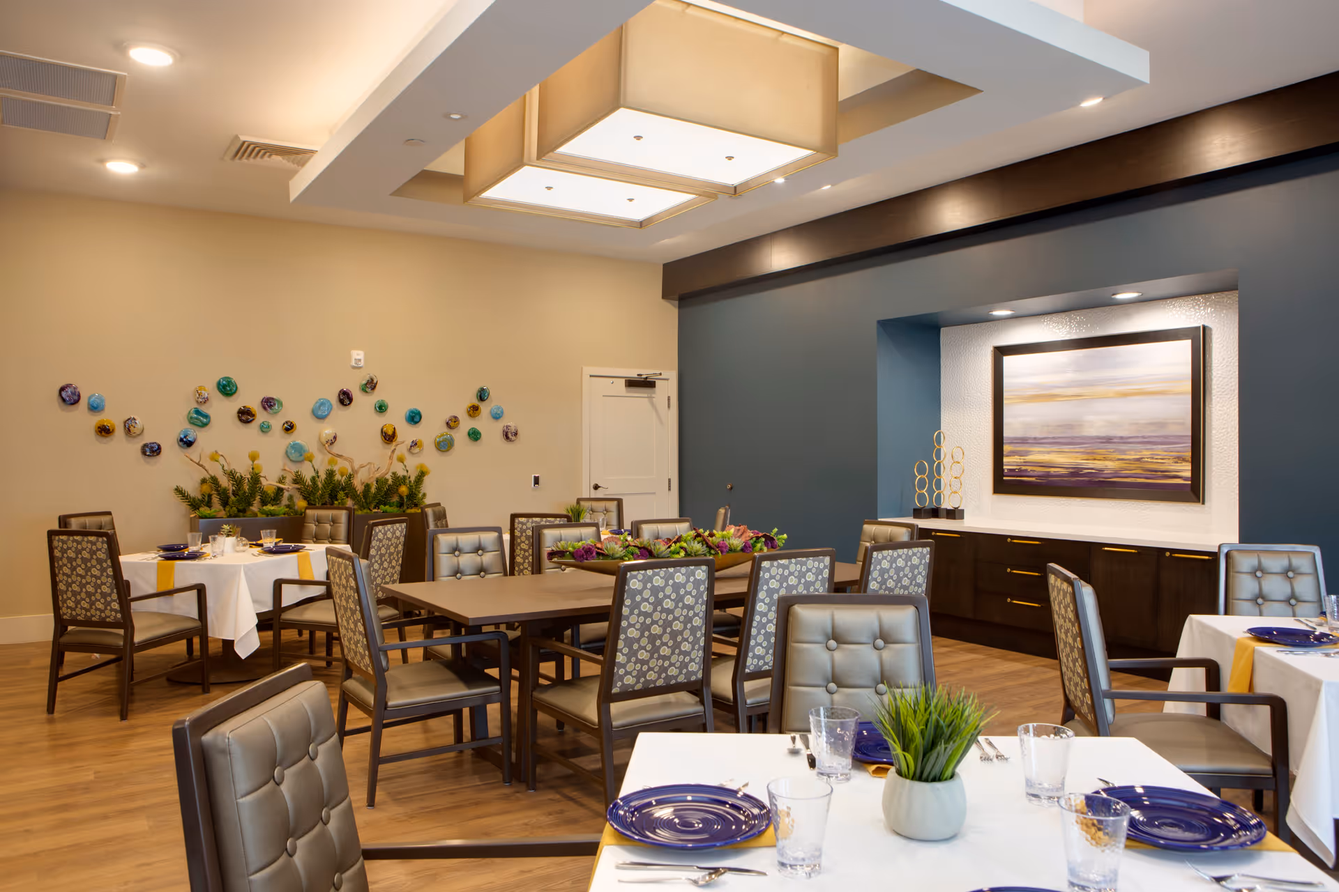 A well-lit dining room with multiple tables set with white tablecloths, blue plates, glasses, and silverware. The room features wooden flooring, cushioned chairs with patterned backs, a decorative wall with colorful circular art pieces, and a large framed painting on a blue accent wall. There are plants and modern ceiling lights adding to the ambiance.