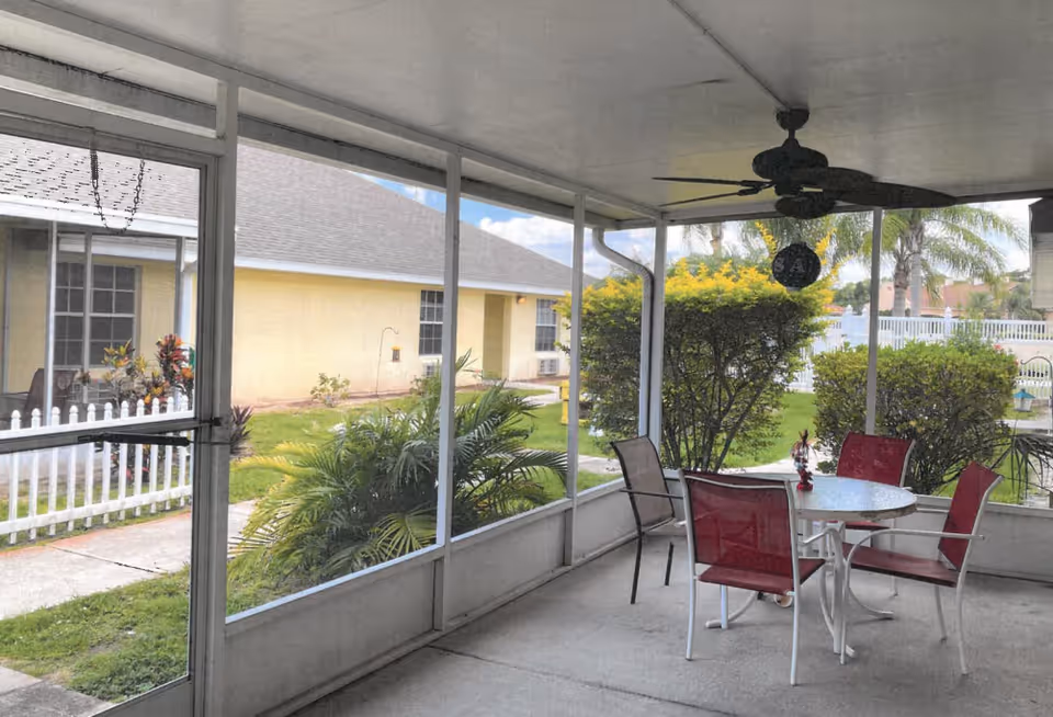 Screened-in patio with a round table and red chairs under a ceiling fan, overlooking shrubs, a white picket fence, and a neighboring building.