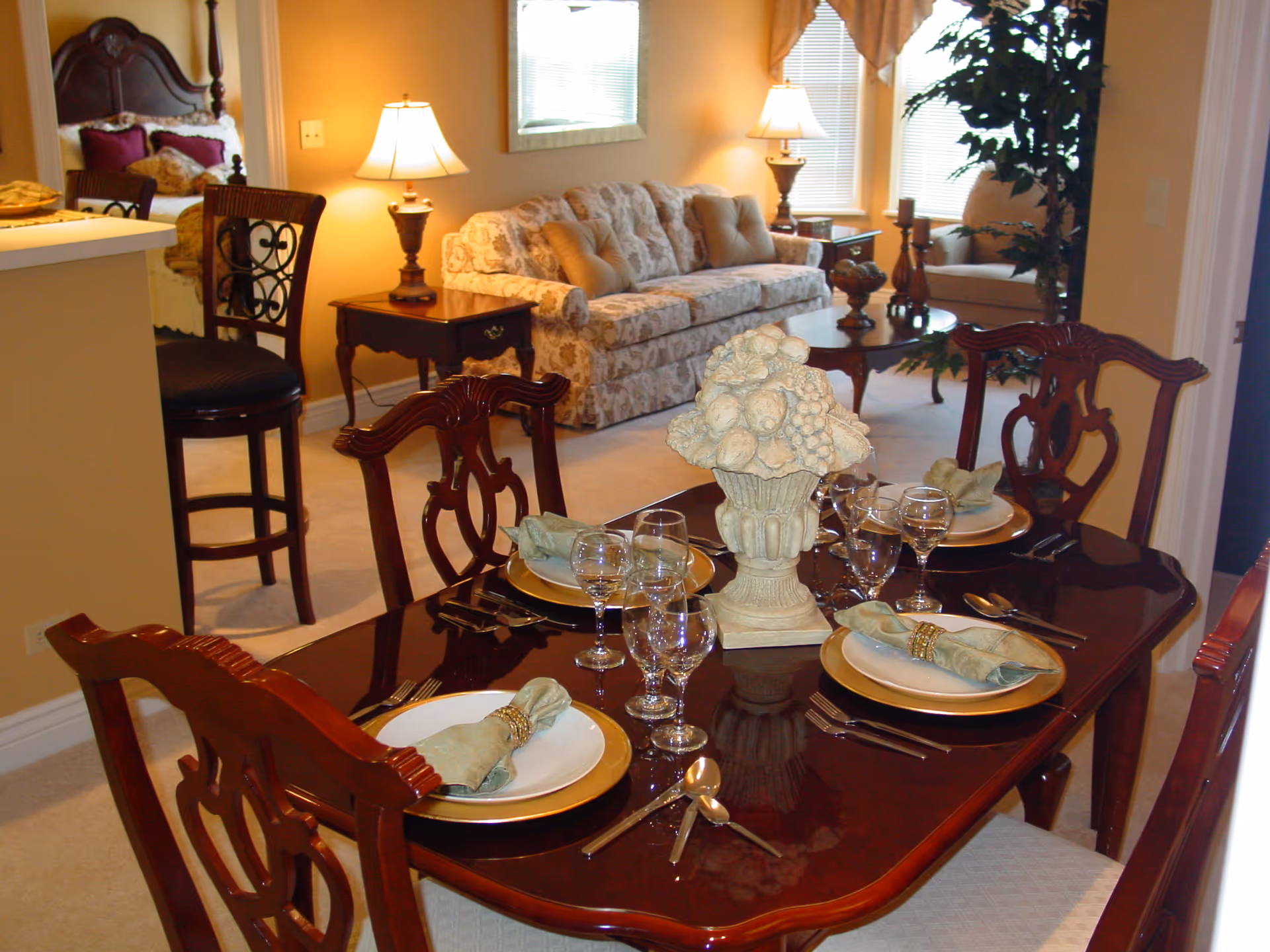 A formal dining table set with plates, glassware and a decorative centerpiece, with a living room and sofa visible in the background.
