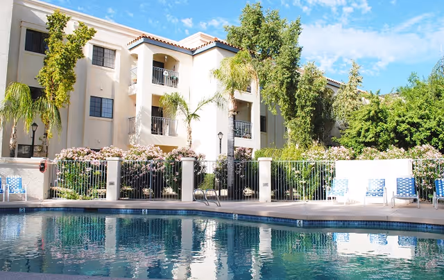 Outdoor swimming pool in front of a Mediterranean-style multi-story senior living building with balconies, palm trees, and lounge chairs.