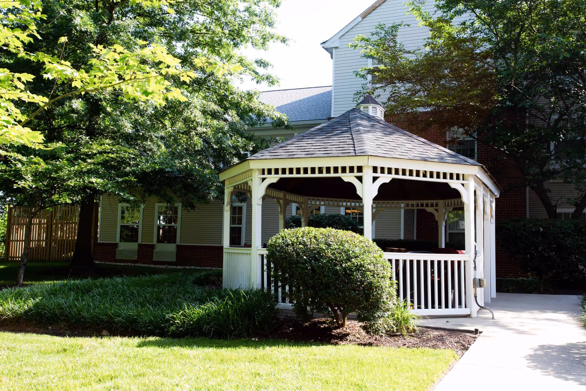 A white wooden gazebo sits on a grassy courtyard in front of a multi-story building surrounded by trees and shrubs.