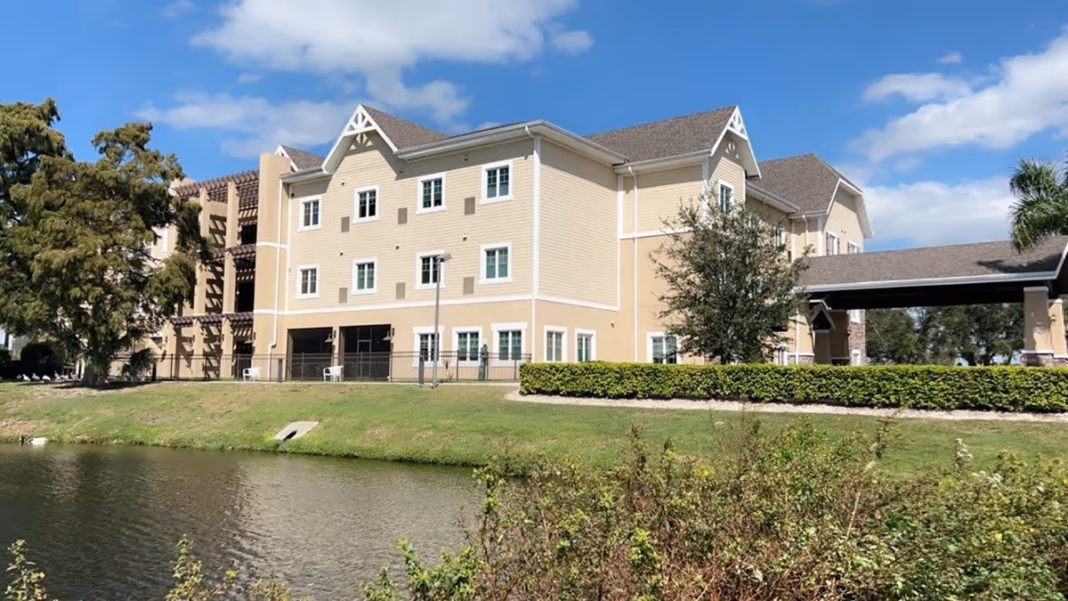 Exterior view of a three-story beige assisted living facility building with white trim, surrounded by greenery and a small body of water in the foreground under a partly cloudy sky.