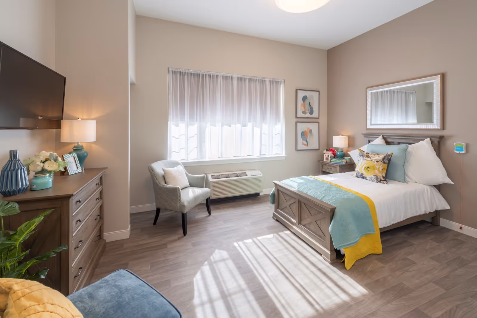 A bright and cozy bedroom with a single bed adorned with white, yellow, and teal bedding and decorative pillows. There is a wooden nightstand with a lamp and flowers beside the bed, a large mirror above the bed, and two abstract art pieces on the wall. A comfortable armchair is placed near a window with sheer white curtains, and a wooden dresser with decorative items and a mounted flat-screen TV is visible. The room has wood flooring and neutral-colored walls.