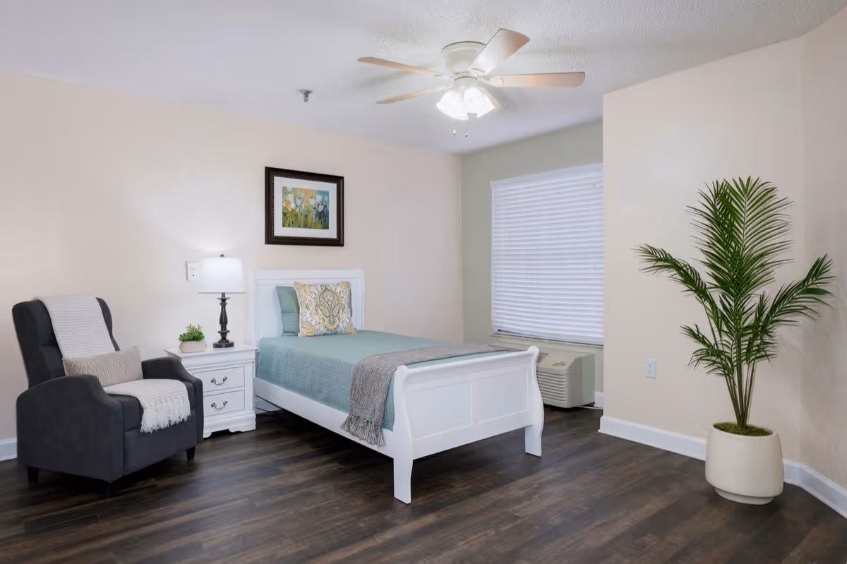 A neatly arranged bedroom with a white single bed featuring a teal bedspread and decorative pillow. Next to the bed is a white nightstand with a lamp and a small plant. A dark gray armchair with a light throw blanket and pillow is positioned beside the nightstand. There is a framed floral painting on the wall above the bed, a ceiling fan with lights, a window with closed blinds, and a tall potted plant in the corner. The room has dark wood flooring and light-colored walls.