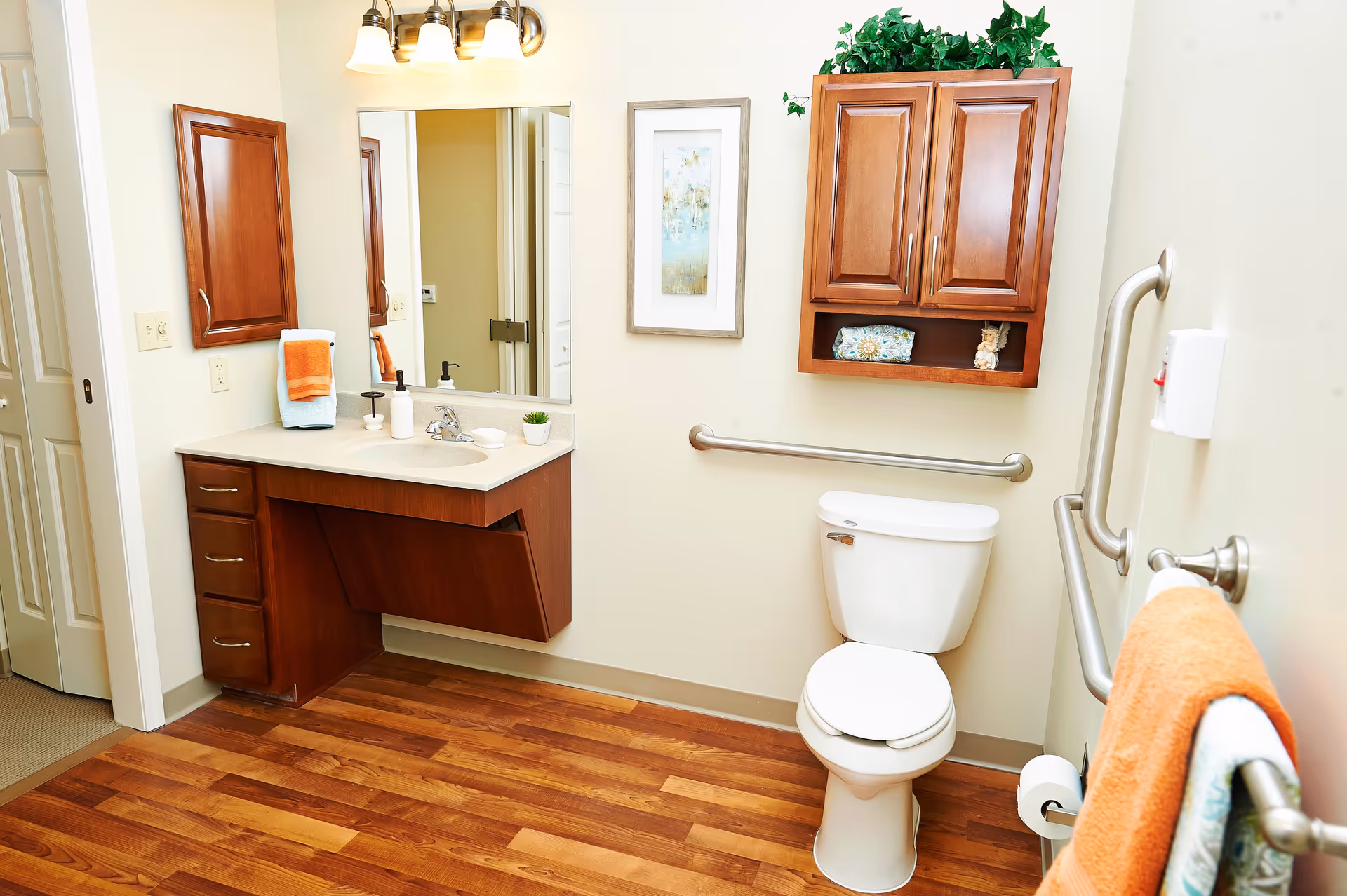 A clean and well-lit bathroom with wooden flooring, a white toilet with grab bars on the walls for support, a wooden vanity with a sink and countertop, a large mirror above the sink, a wooden cabinet mounted on the wall with decorative items and greenery on top, and orange and blue towels hanging on a towel rack.