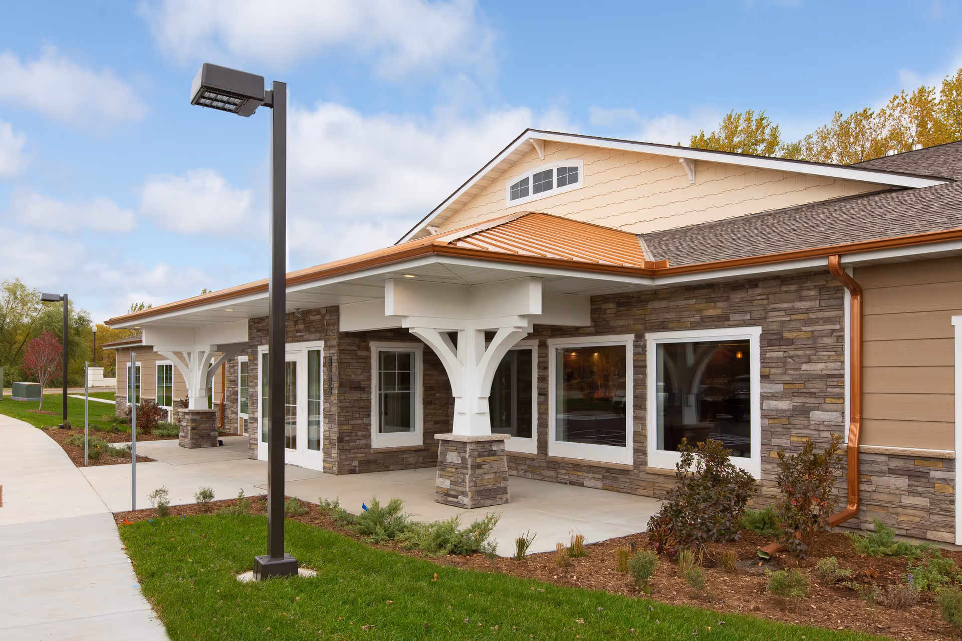 Exterior front entrance of a single-story senior living building with a covered portico, stone facade, large windows, and landscaped grounds.