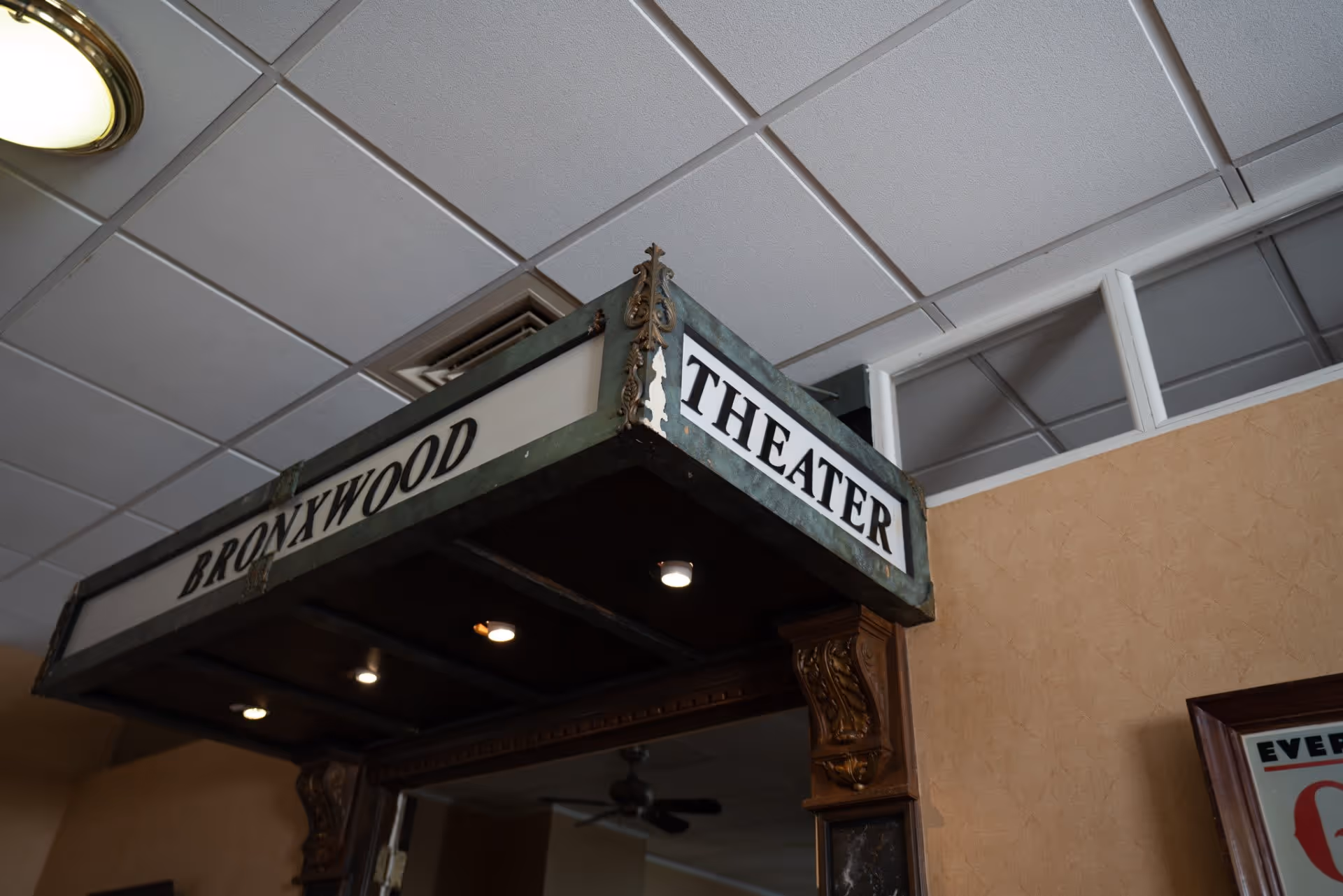Interior view of an entrance to the Bronxwood Theater, featuring a vintage-style marquee sign with the words 'BRONXWOOD THEATER' in black letters on a white background. The ceiling has white tiles and recessed lighting is visible under the marquee.