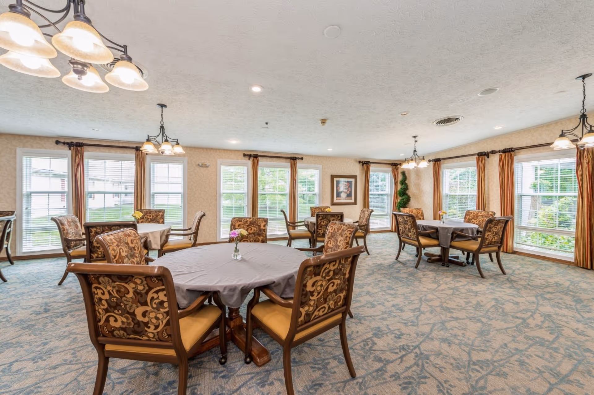 A bright dining room with multiple round tables covered with tablecloths and surrounded by patterned chairs. Large windows with curtains allow natural light to fill the room. Ceiling lights hang above the tables, and a framed picture and a tall plant decorate the walls.