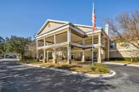 Front entrance of a two-story yellow senior living building with a covered portico, American flag on a flagpole, and a circular driveway.