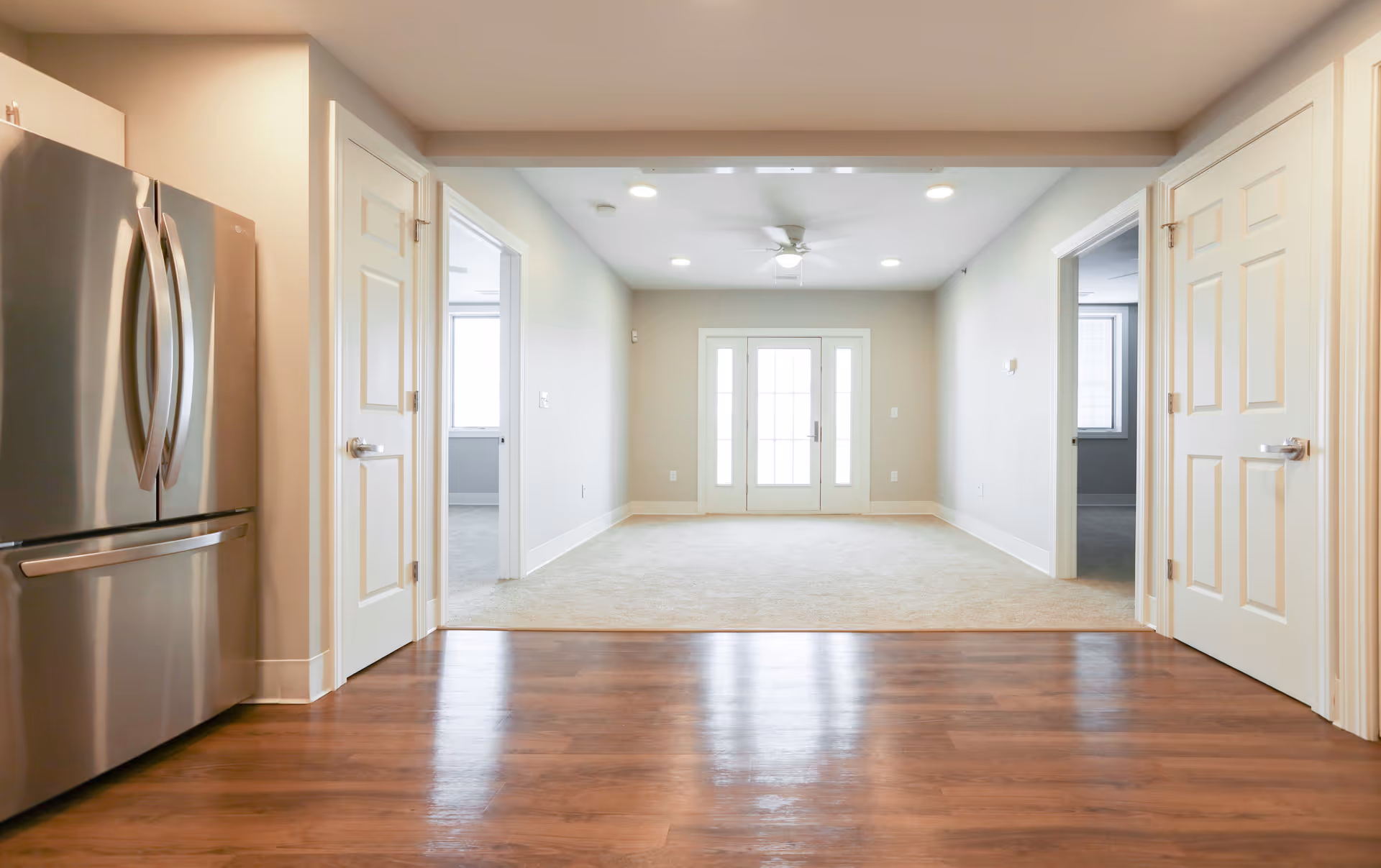 Interior view of a modern apartment with wooden flooring in the foreground leading to a carpeted living area. The living area has a ceiling fan with lights and a set of glass double doors at the far end. On either side of the living area are doorways leading to other rooms. A stainless steel refrigerator is visible on the left side of the image.