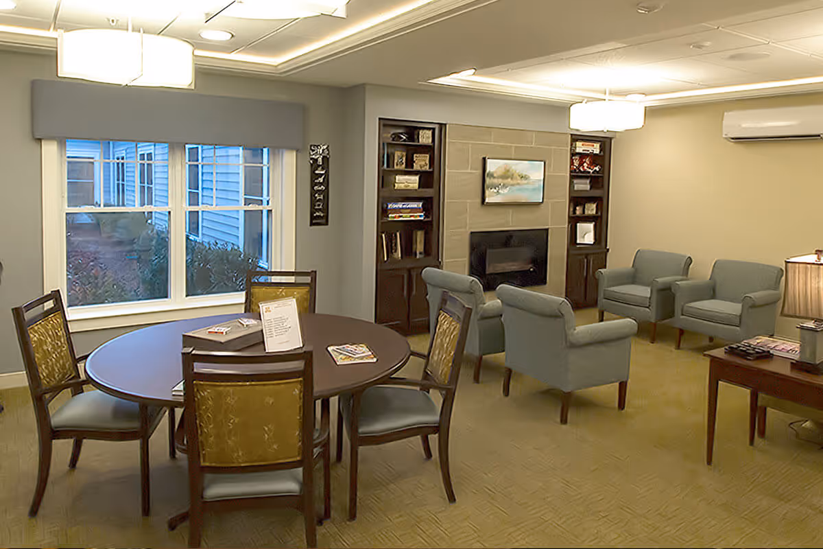 A cozy living room area with a round wooden table surrounded by four chairs with green upholstery. In the background, there are four light blue armchairs arranged around a fireplace with a painting above it. Built-in shelves flank the fireplace, holding books and decorative items. A large window shows an outdoor view of a building and bushes. The room is softly lit with ceiling lights and a table lamp.