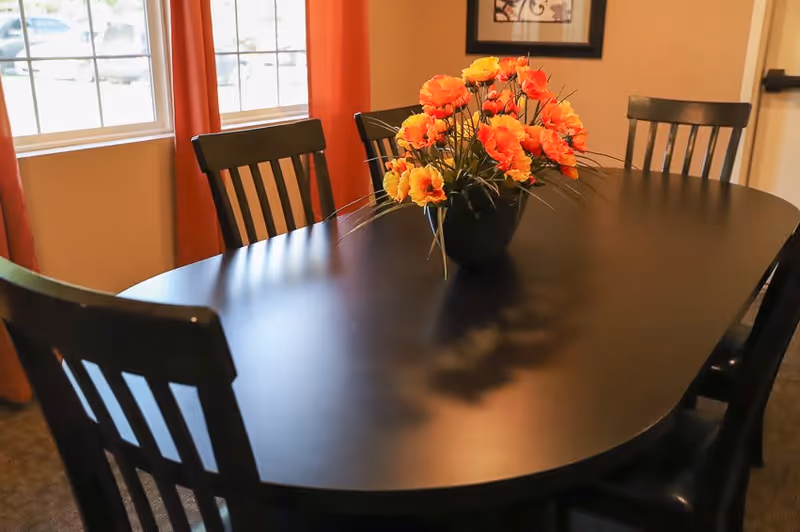 A dark wooden dining table with six matching chairs around it, featuring a centerpiece of bright orange and yellow flowers in a black vase. The room has beige walls, a window with red curtains, and a framed picture on the wall.