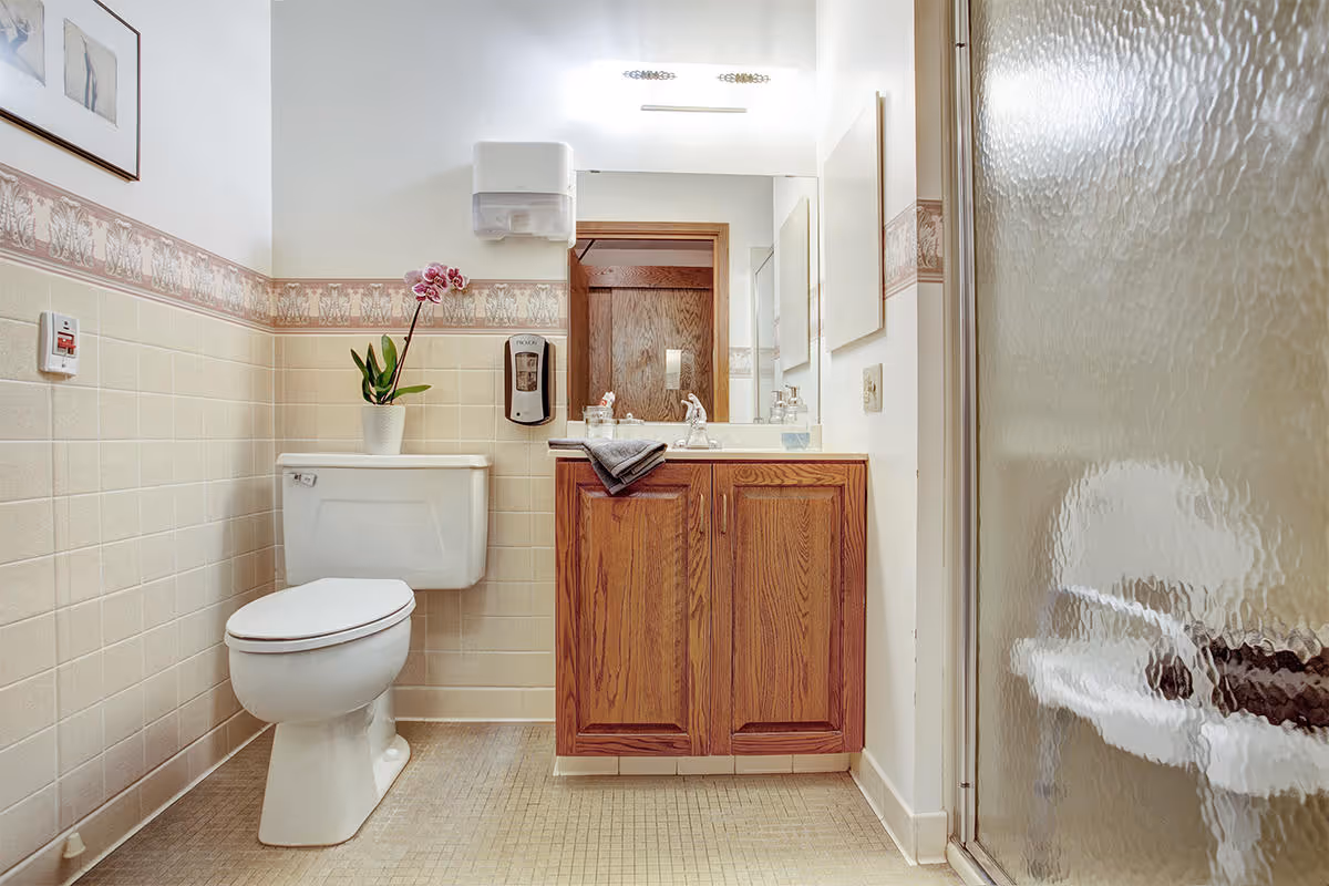 A bathroom with a white toilet, a wooden vanity with a sink, a mirror above the sink, a wall-mounted soap dispenser, a paper towel dispenser, and a shower with a textured glass door. There is a potted orchid plant on top of the toilet tank and a framed picture on the wall.