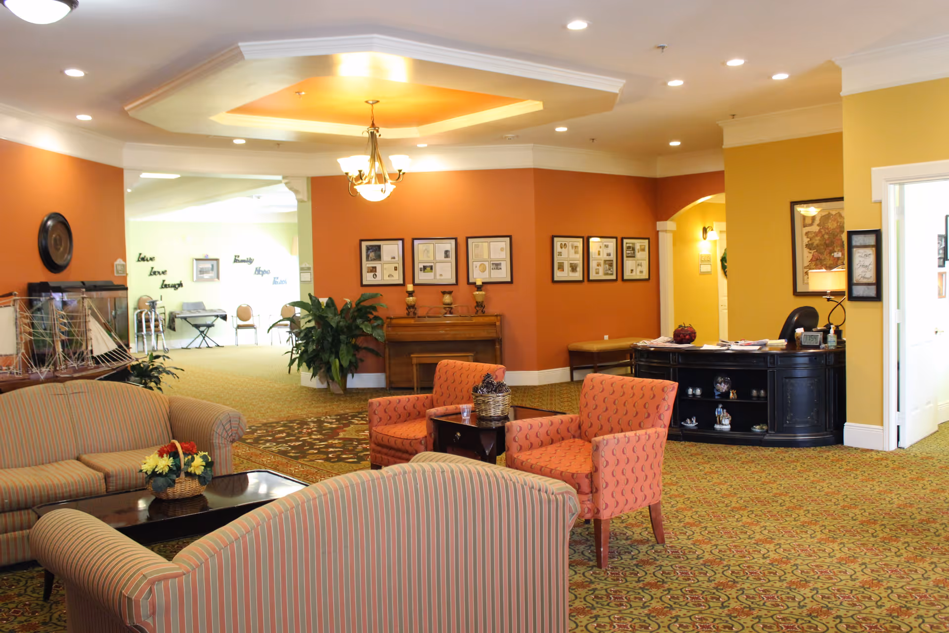 A cozy senior living facility common area with striped sofas, two orange patterned armchairs, a coffee table with a flower basket, and a piano against an orange wall. The room has warm yellow and orange walls, framed pictures, a chandelier, and a patterned carpet. In the background, there is an open space with chairs and a keyboard, and motivational words on the wall.