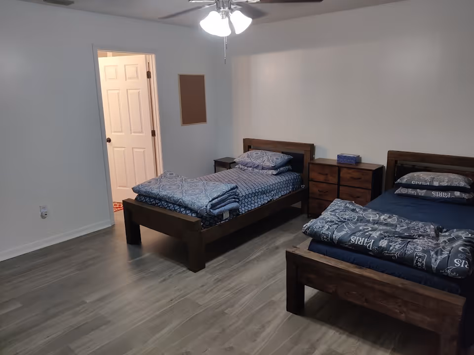 Sparsely furnished bedroom with two twin wooden beds, a chest of drawers between them, and a ceiling fan.