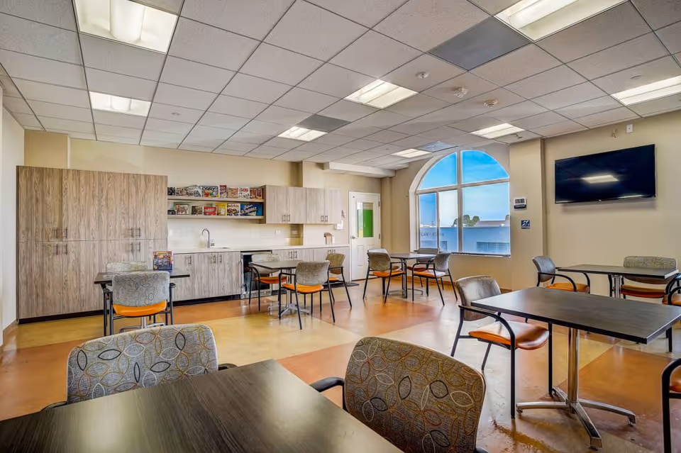 Bright communal dining/activity room with tables and chairs, a kitchenette with cabinets, and a wall-mounted TV by a large arched window.