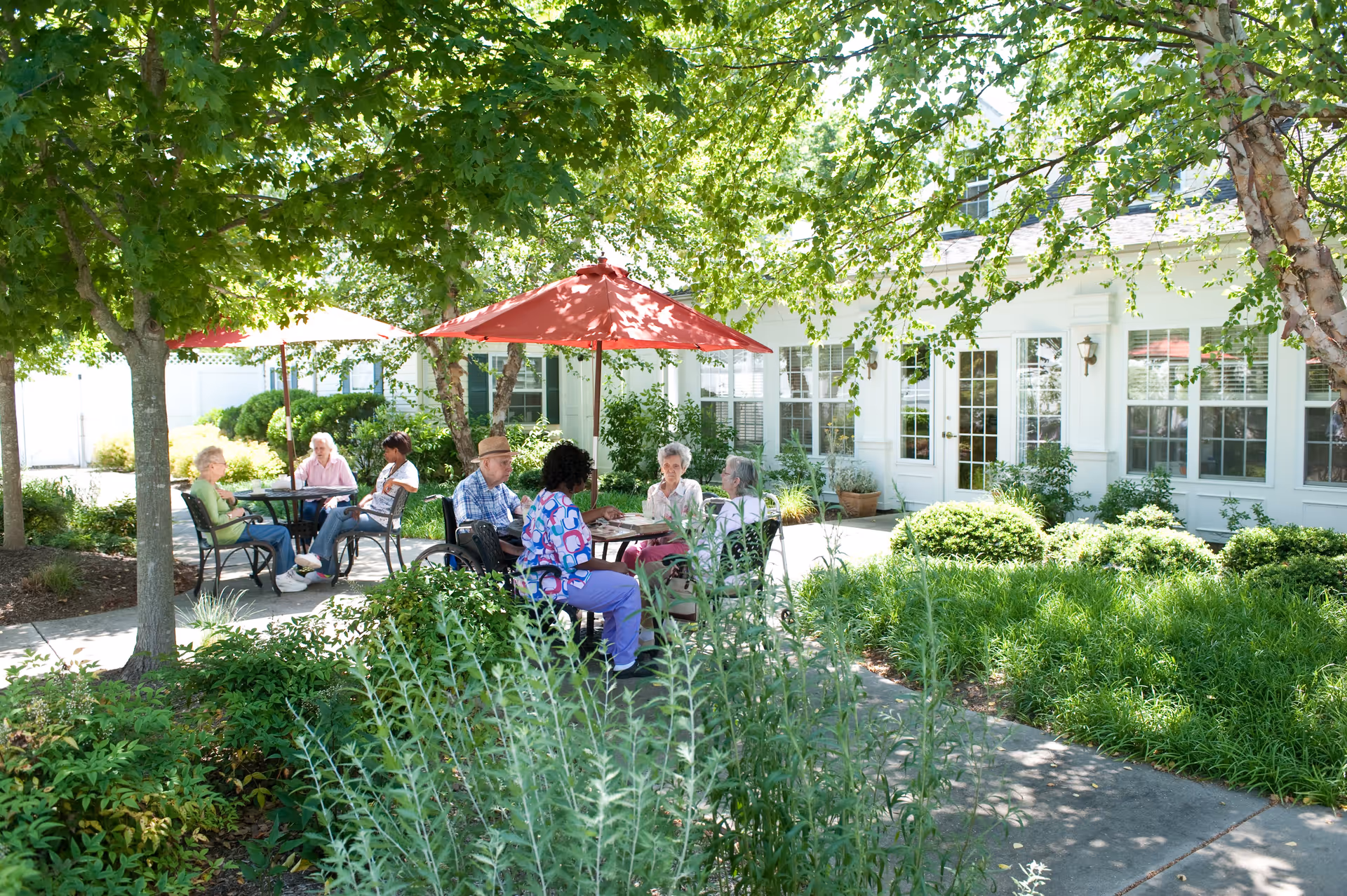A group of elderly people sitting at outdoor tables under red umbrellas in a garden area with trees and greenery, next to a white building with large windows and doors.