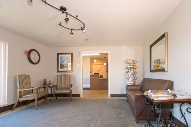 A waiting area with two striped armchairs and a small table with flowers on the left, a brown leather couch on the right with a side table holding magazines, a large mirror above the couch, a clock and framed picture on the far wall, and an open doorway leading to a reception desk area.