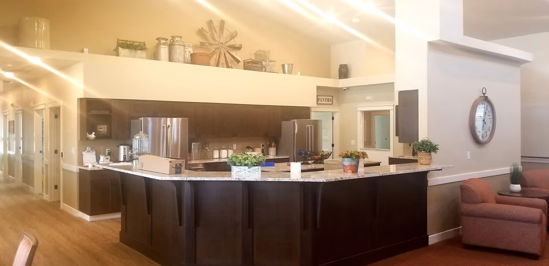 Interior view of a senior care facility kitchen area with dark wood cabinets, stainless steel refrigerator, and a large granite countertop island. The kitchen is decorated with plants and small decorative items on the counter and shelves above the cabinets. To the right, there is a seating area with two upholstered chairs and a large wall clock.