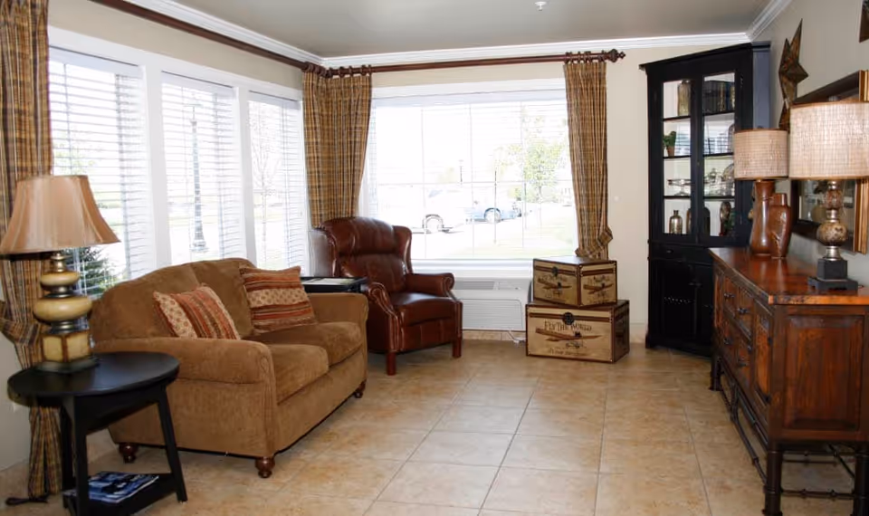 A cozy living room with a brown upholstered loveseat with patterned cushions, a dark brown leather armchair, and a black round side table with a lamp. There are large windows with plaid curtains letting in natural light. Against the wall is a wooden sideboard with two lamps and decorative items, and a black glass-front cabinet. Two vintage-style wooden trunks are stacked near the window.
