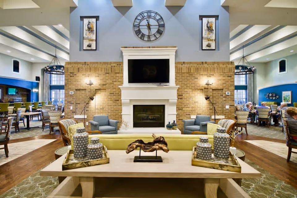 A spacious and well-lit common area in a senior living facility featuring a central fireplace with a mounted TV above it, flanked by two armchairs. In front of the fireplace is a wooden table with decorative items and a green sofa. The room has high ceilings with recessed lighting and large windows. To the left and right, there are dining areas with tables and chairs where several seniors are seated and conversing.