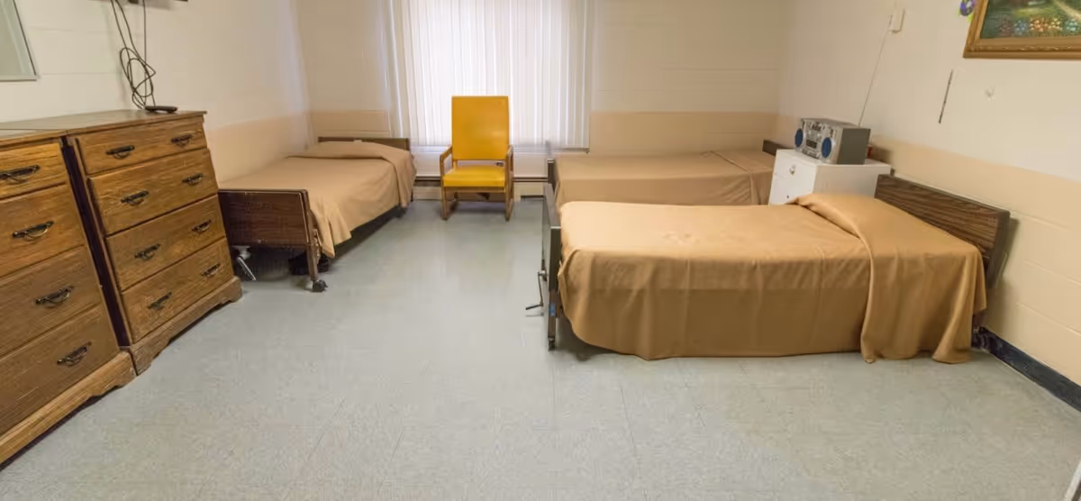 A simple room with three single beds covered in beige bedspreads, a wooden dresser on the left, a yellow chair in front of a window with vertical blinds, and a small white cabinet with a radio on top. The walls are painted white with a beige stripe, and there is a framed picture hanging on the right wall.